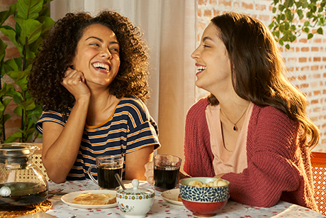 Duas mulheres sentadas em uma mesa com comidas e bebidas, tomando café da marca Café Damasco.